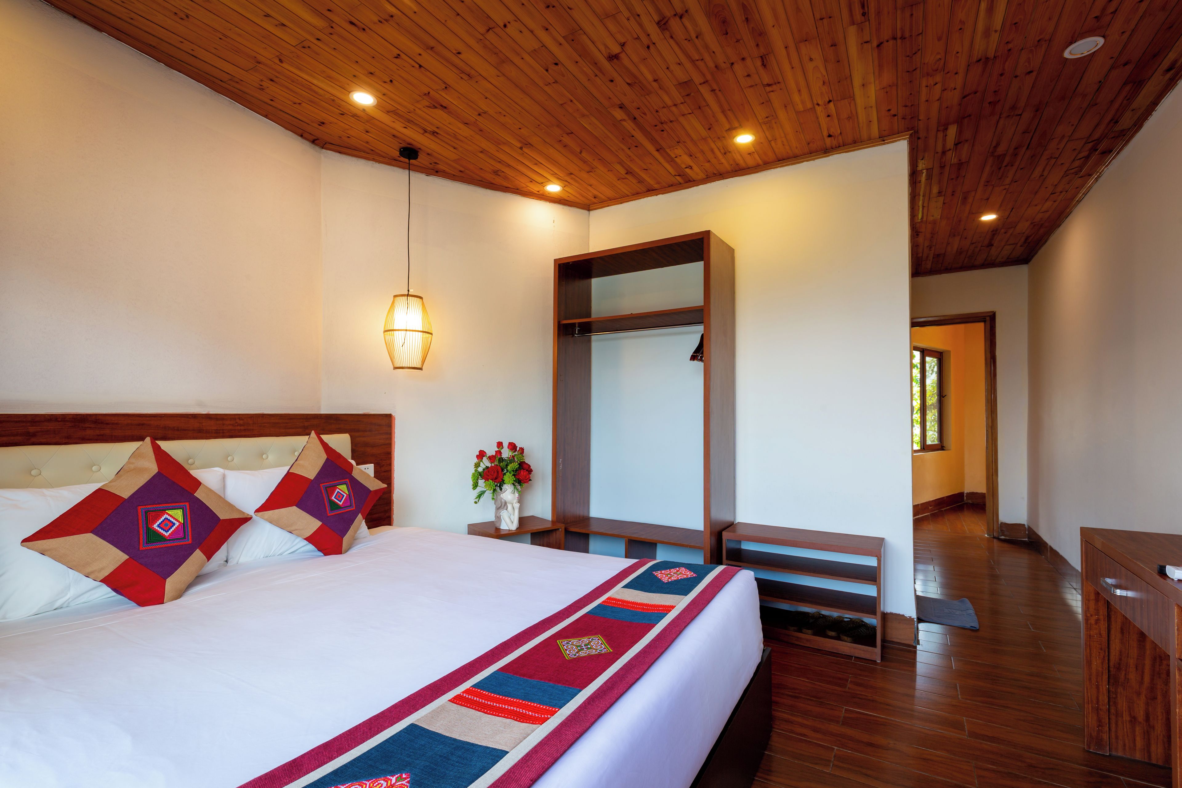 A hotel room with a large bed, a wooden ceiling, a nightstand with a vase of flowers, and a mirror.