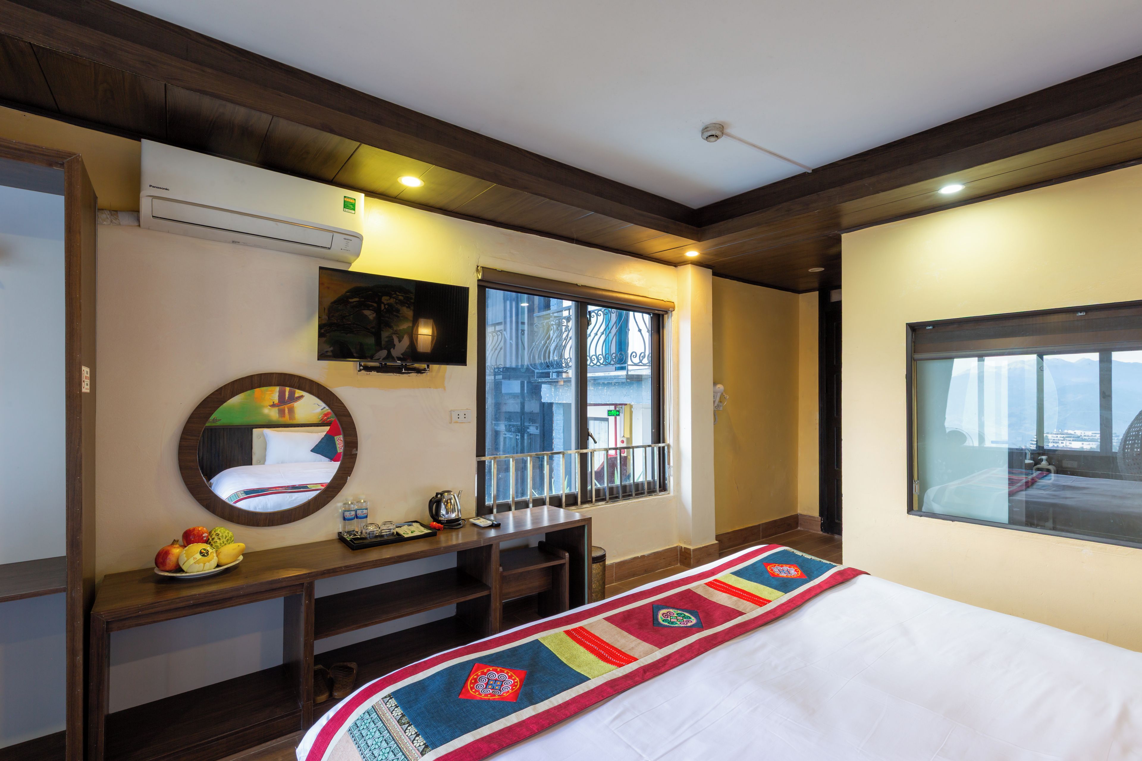 Quarto de hotel com cama, escrivaninha com chaleira, espelho e janela com vista para o exterior.
