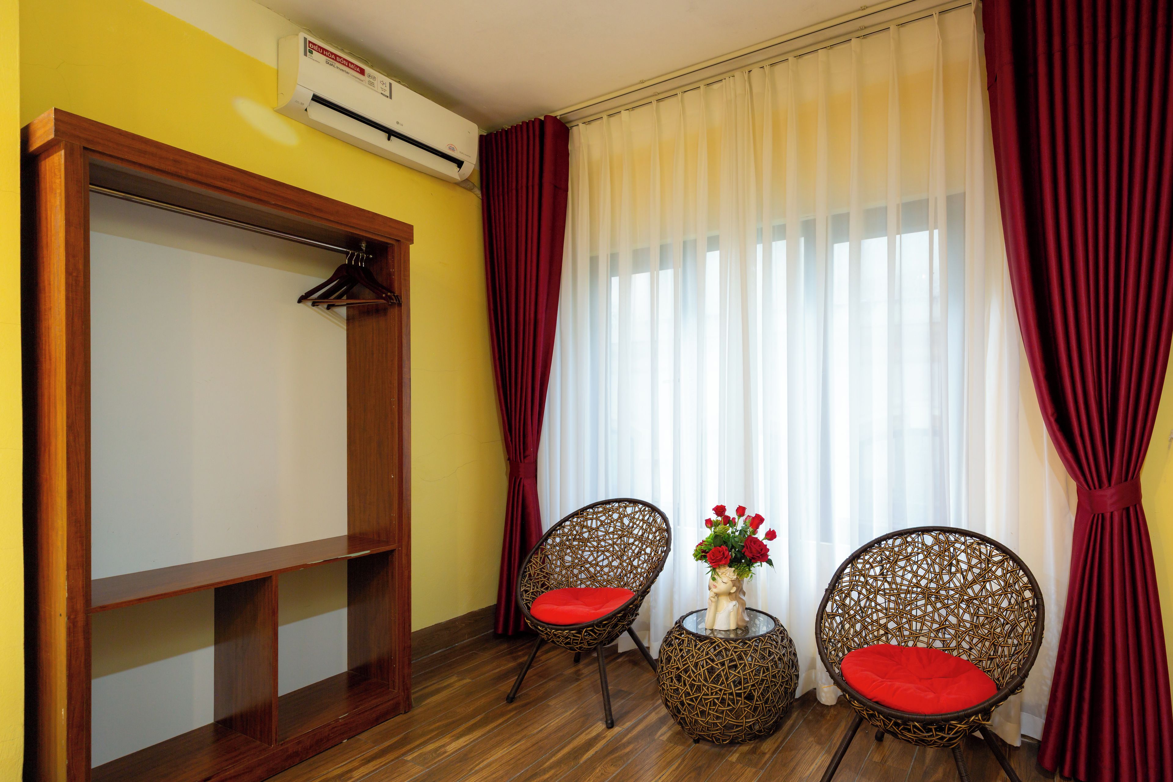 Una habitación con un armario de madera, dos sillas de mimbre con cojines rojos, una mesita con un florero y una ventana con cortinas blancas traslúcidas y cortinas rojas.