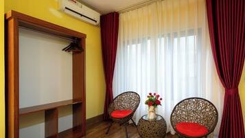 Una habitación con un armario de madera, dos sillas de mimbre con cojines rojos, una mesita con un florero y una ventana con cortinas blancas traslúcidas y cortinas rojas.