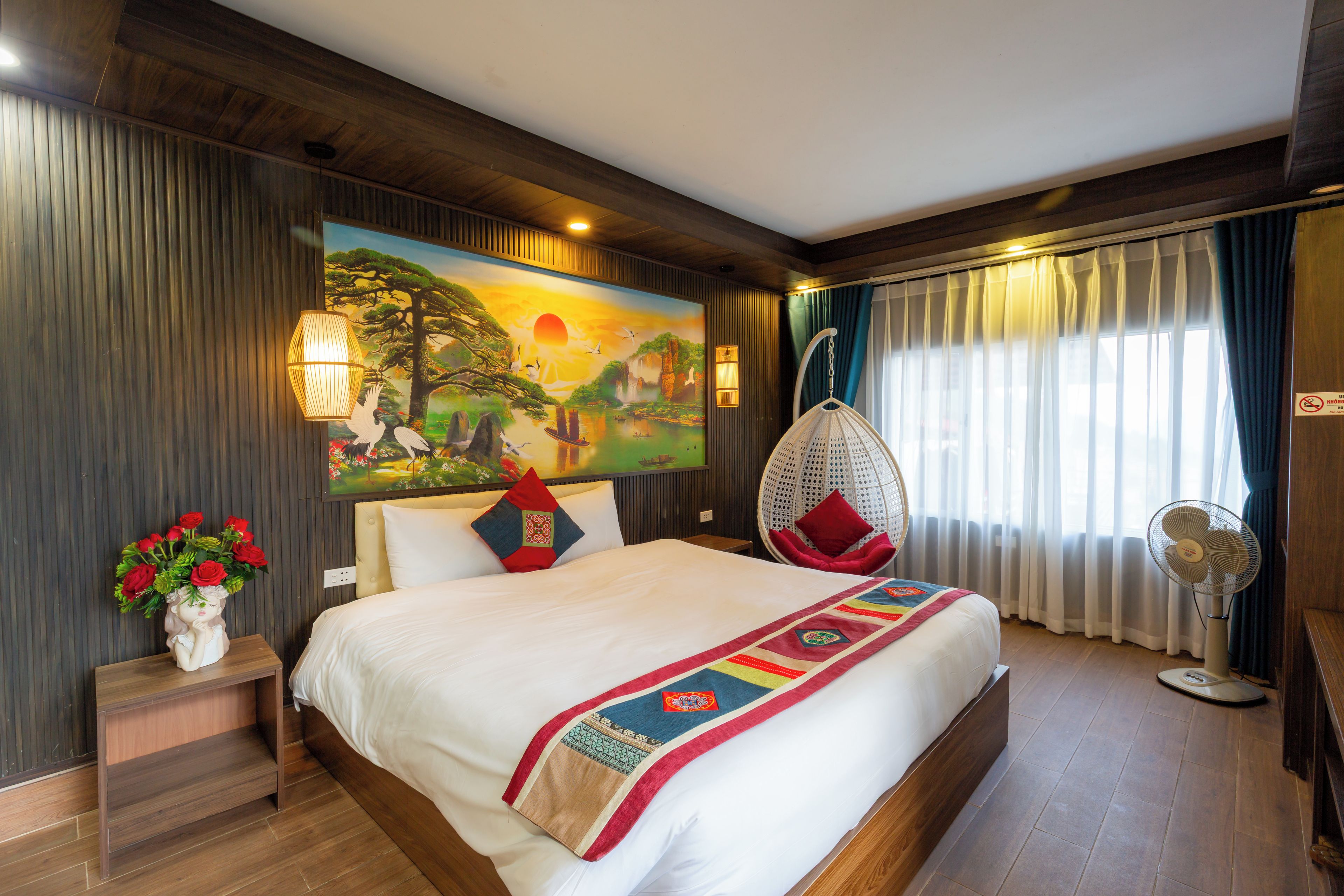 A hotel room with a bed, a hanging chair, a wall painting, and a window with curtains.