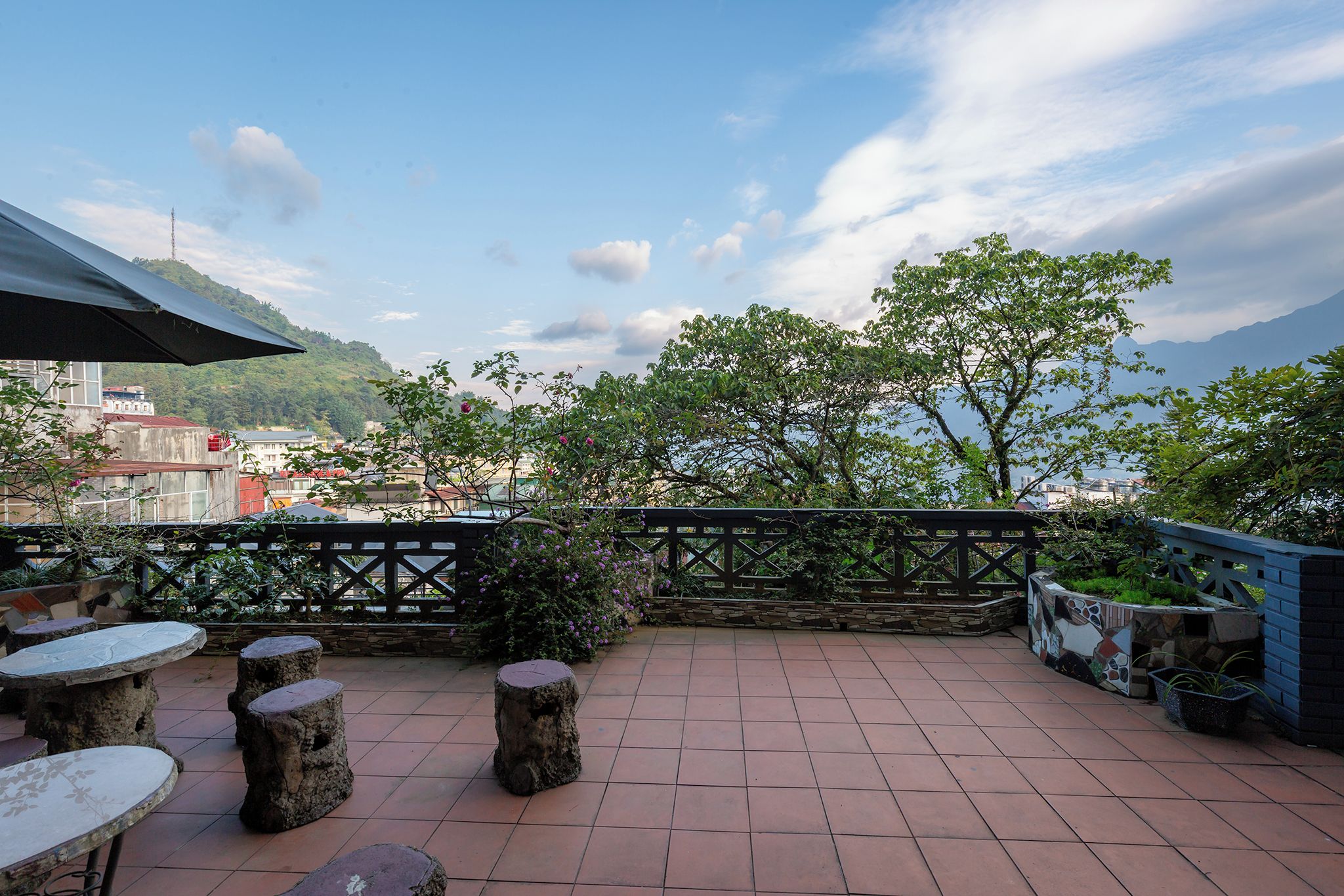 Um pátio externo com mesas e bancos de pedra, cerca decorativa e vista para um horizonte urbano e montanhas ao fundo.