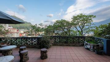 Um pátio externo com mesas e bancos de pedra, cerca decorativa e vista para um horizonte urbano e montanhas ao fundo.