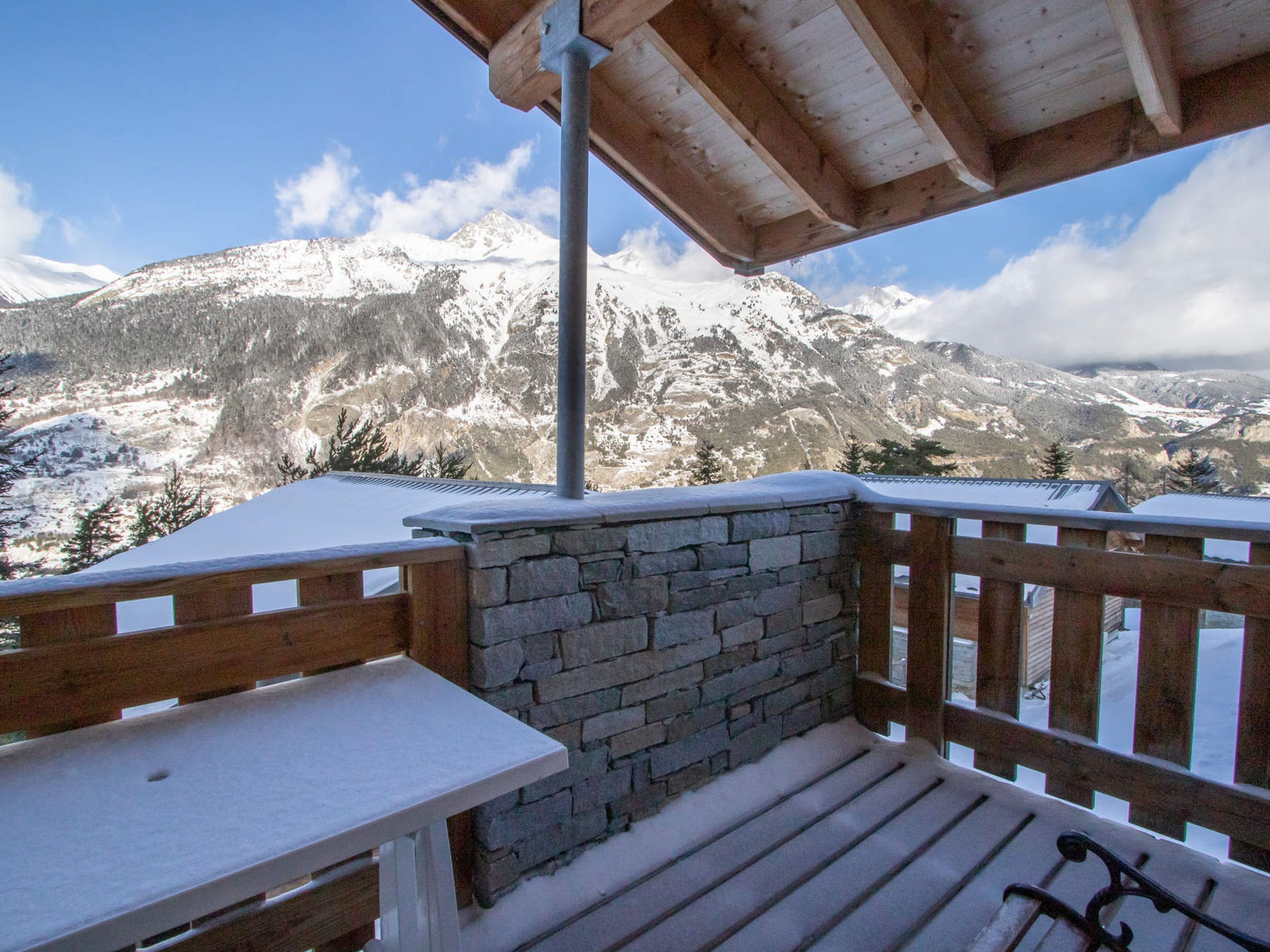 Una terrazza in legno con un muro in pietra e un tavolo, con vista su una catena montuosa innevata.