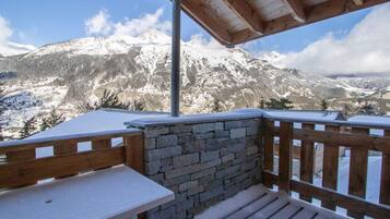 Una terrazza in legno con un muro in pietra e un tavolo, con vista su una catena montuosa innevata.