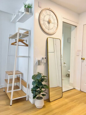 A room with a wooden floor, a white wall-mounted clock, a mirror, a plant, a white shelf with decorative items, and a door leading to a bathroom. - Aloha Bliss Surfers Paradise Beach Stay (Surfers Paradise)