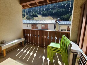 Ein Balkon mit Blick auf eine bewaldete Berglandschaft, einer Holzbank und grünen Plastikstühlen.