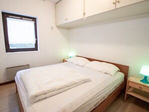 Ein Einzelbett mit Holz Kopfteil, ein Nachttisch mit Lampe, ein Fenster mit Jalousien und ein an der Wand befestigtes Schränkchen.