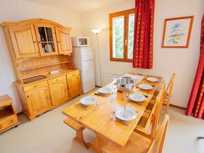 Eine kompakte Küche mit Holzschränken, einer Mikrowelle und einem Kühlschrank. Ein Esstisch ist gedeckt mit Tellern, Besteck und einem großen Topf.