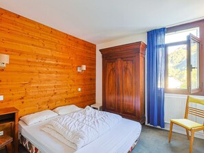 Huoneessa on puinen seinä, valkoisilla lakanoiden peitetty sänky, puinen vaatekaappi, tuoli ja sinisillä verhoilla varustettu ikkuna.