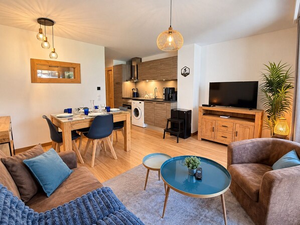 Uma sala de estar moderna com sofá, mesa de centro e televisão. Há uma área de jantar com cadeiras e mesa, e uma cozinha com eletrodomésticos.