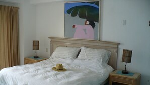 A bedroom with a bed, two bedside tables, a hat on the bed, a painting on the wall, and a curtain. - Loaded 2-bedroom view condo on the prettiest
beach in Vallarta (Jal.)