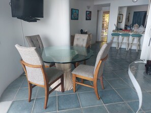 A dining area with a glass-top table surrounded by upholstered chairs, a television mounted on the wall, and a view of the outdoor area through an open door. - Loaded 2-bedroom view condo on the prettiest 
beach in Vallarta (Jal.)