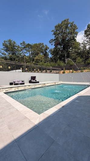 A rectangular swimming pool with a stone edge, surrounded by a tiled deck. There are two lounge chairs and a small table with a canopy. - Entire home in Aldie, Virginia, United States (Aldie)