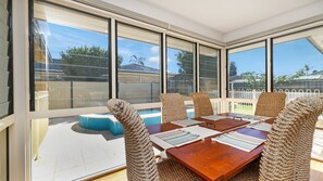 A sunlit patio with a wooden table and woven chairs, large windows, and a pool. - Salt Gardens – Private Pool & Tea Gardens Retreat (Tea Gardens)