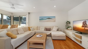 A modern living room with a sectional sofa, a coffee table, a flat-screen TV, and a painting of a beach scene. - Salt Gardens – Private Pool & Tea Gardens Retreat (Tea Gardens)