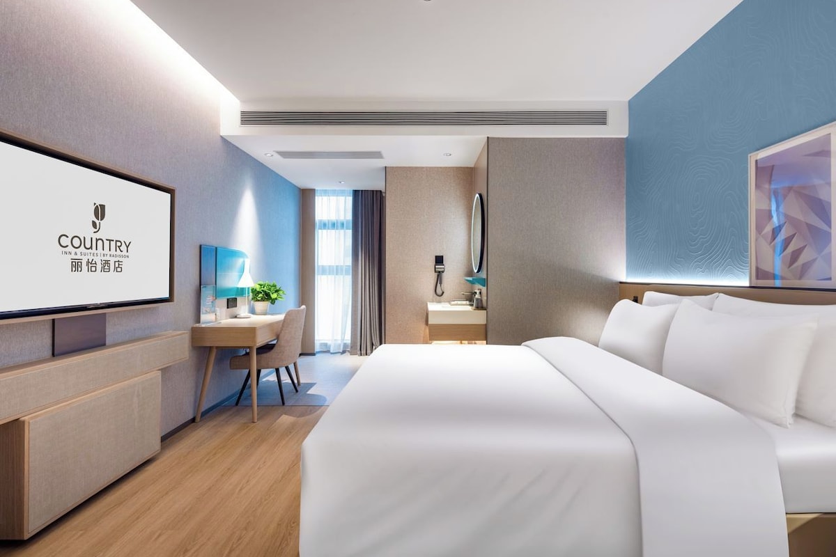 A modern hotel room with a large bed, a desk, and a television displaying the hotel name.