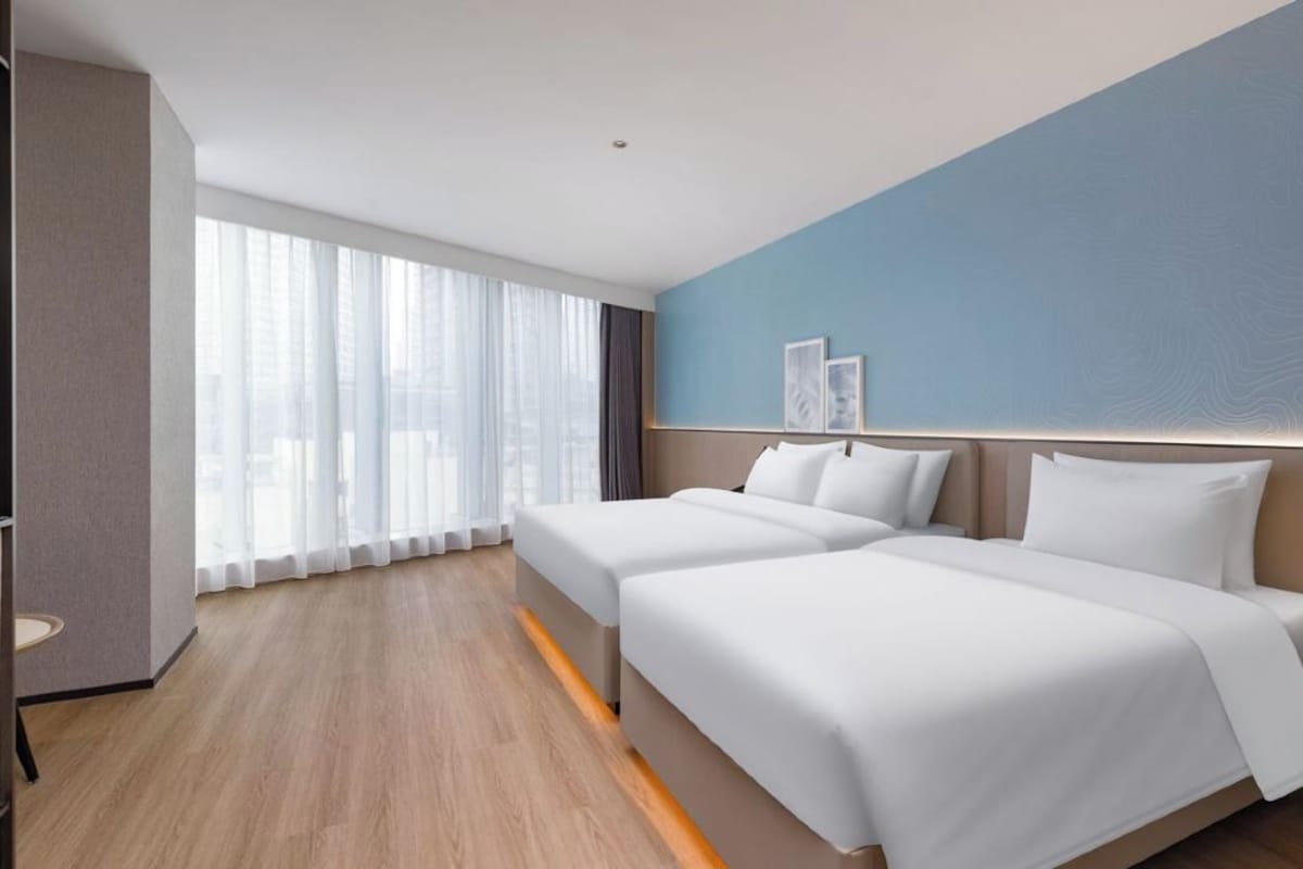 Quarto de hotel com duas camas, uma janela grande com cortinas, parede azul de destaque e uma TV de tela plana.