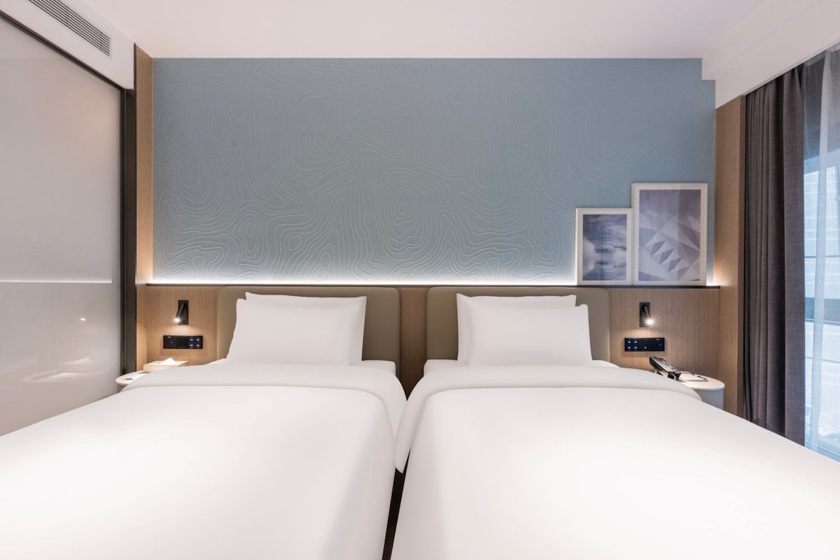 Quarto de hotel com duas camas, cabeceira moderna, mesas de cabeceira com luminárias e obra de arte na parede.