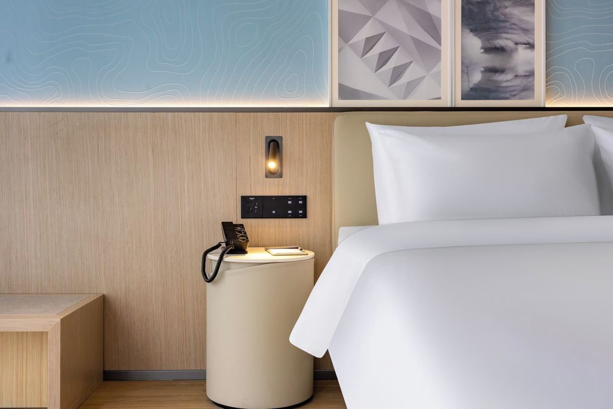 A modern hotel room with a wooden headboard, a bedside table with a lamp, and artwork on the wall.