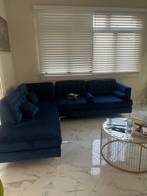 A living room with a dark blue sectional sofa, a glass coffee table, and a window with blinds. - The Chesterfield House: Luxury Caribbean Escape in Barbados (St. Michael, M10)