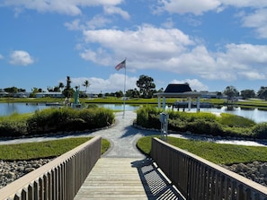 A wooden boardwalk leading to a gazebo over a pond with an American flag, surrounded by greenery and a clear sky.