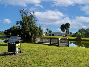 A grassy area with a grill, a wooden dock over water, and a house in the background.
