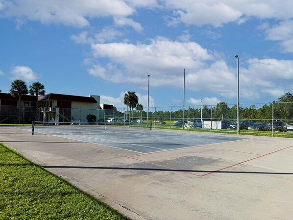 An outdoor tennis court with a net, surrounded by a fence, and a building in the background.