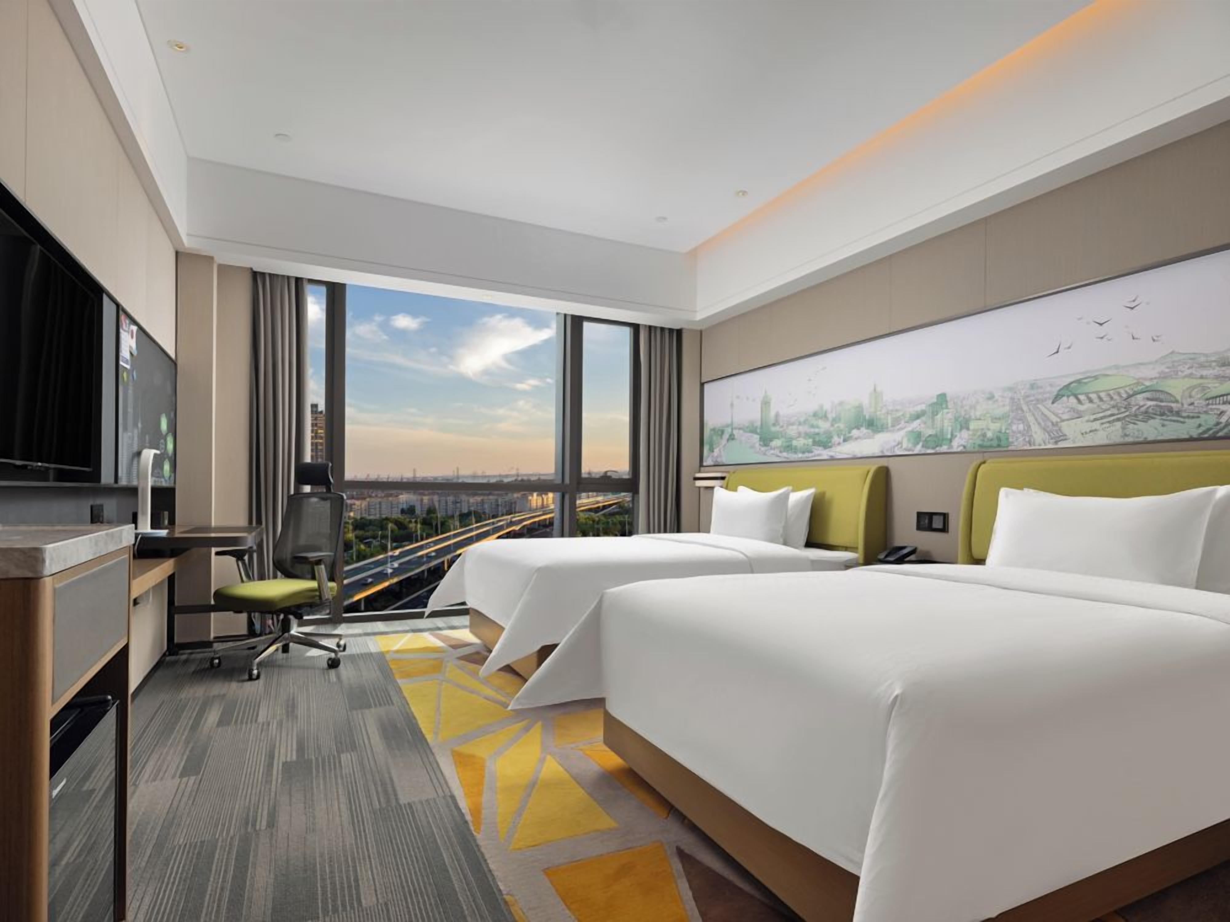 A hotel room with two beds, a desk, a chair, and a large window offering a city view.