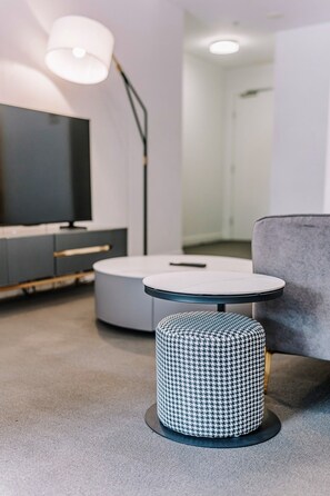 Ein modernes Wohnzimmer mit einem runden weißen Tisch, einem Hunderzahn-Pouff, einem grauen Sofa und einer Stehlampe.