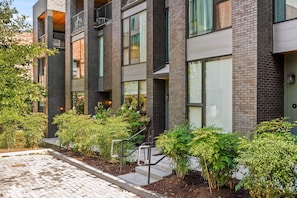 Een modern meerlaags gebouw met een bakstenen gevel, grote ramen en een aangelegde tuin met struiken en een kleine trap naar de ingang.