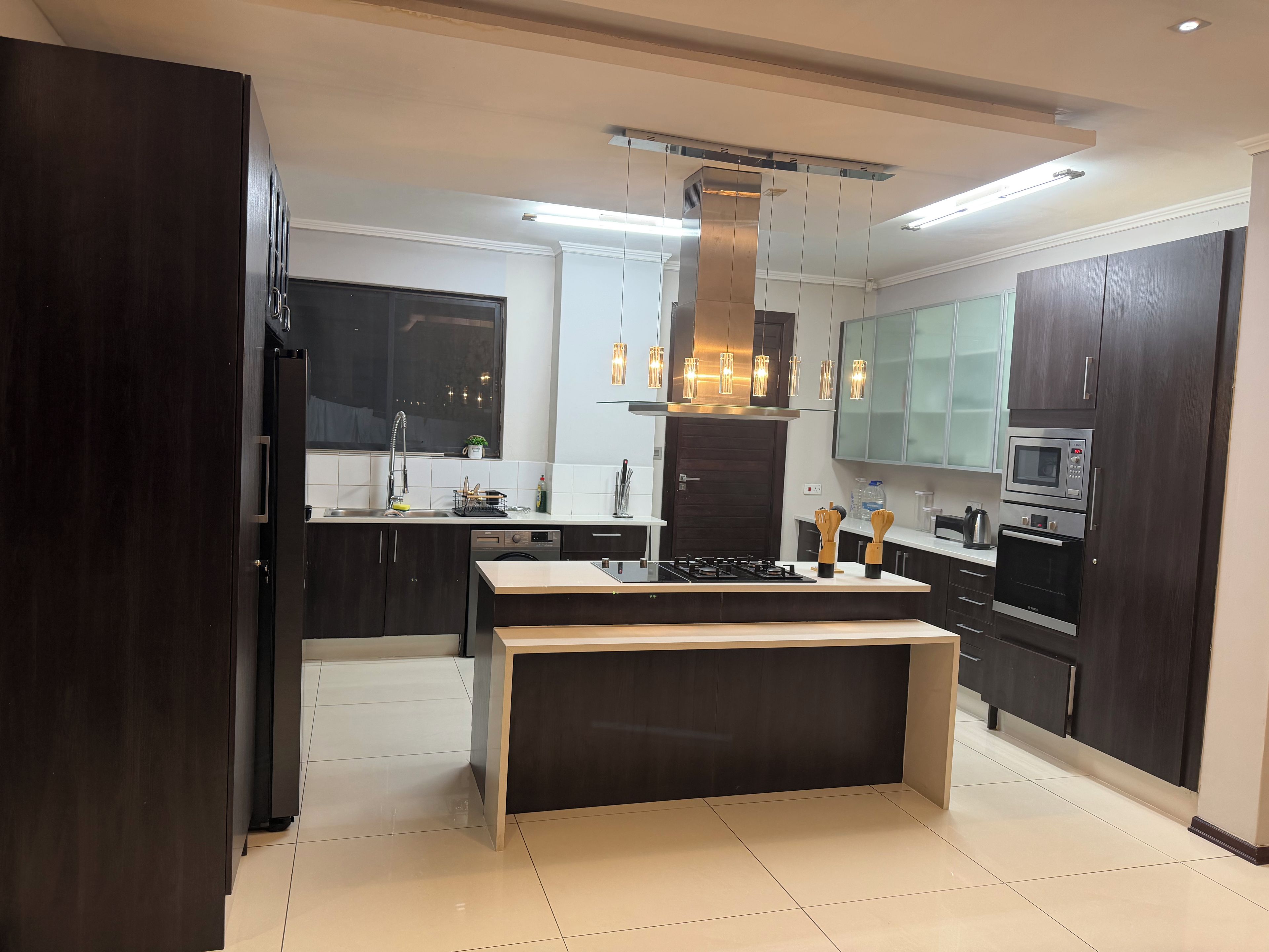 Une cuisine moderne avec des meubles de couleur sombre, un îlot central, des appareils électroménagers en acier inoxydable et des luminaires suspendus.