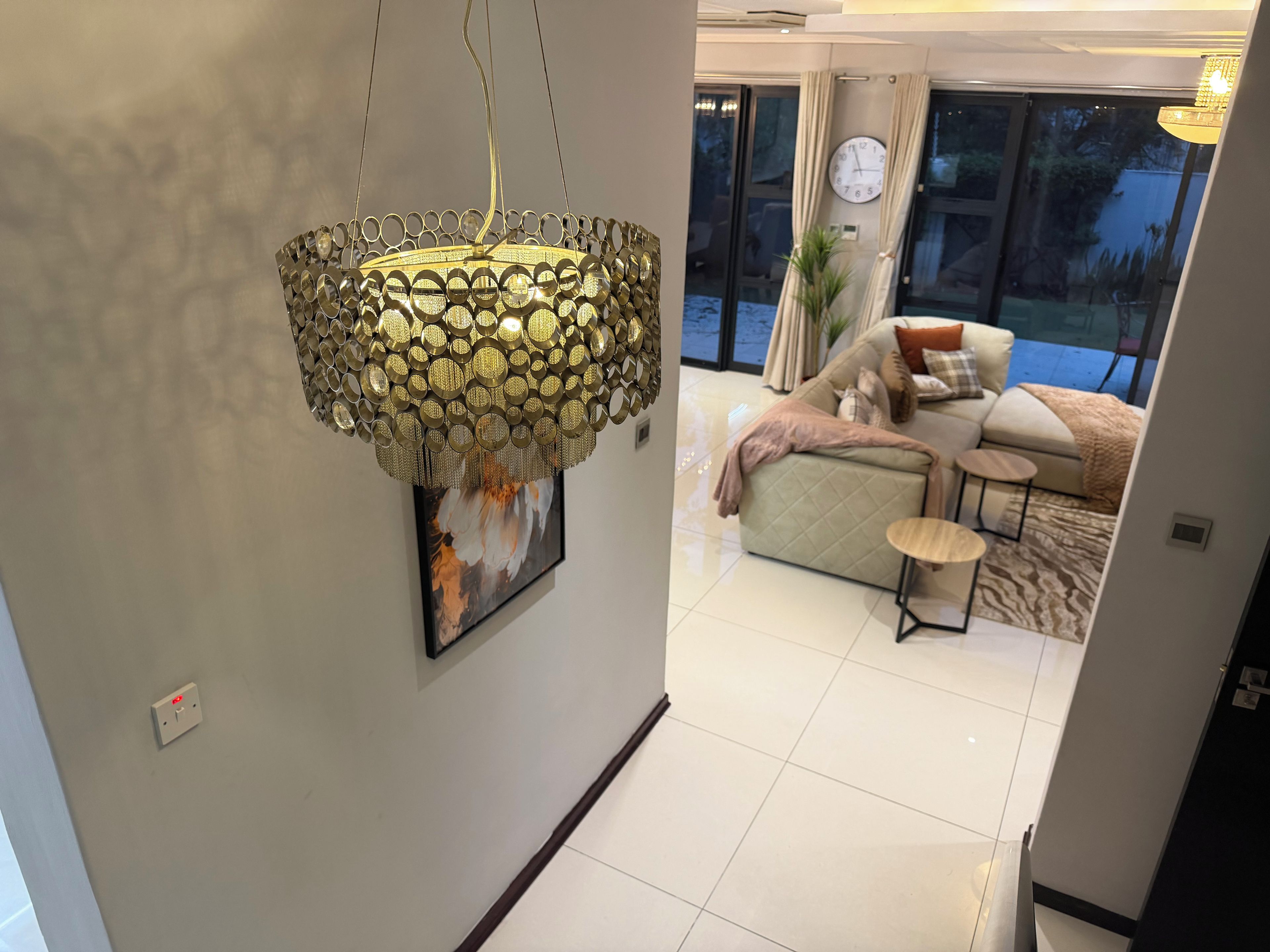 Un intérieur moderne agrémenté d’un luminaire décoratif, d’une œuvre d’art encadrée et d’un espace de vie comprenant un canapé, une table basse et une vue sur l’extérieur, visible à travers une porte vitrée.