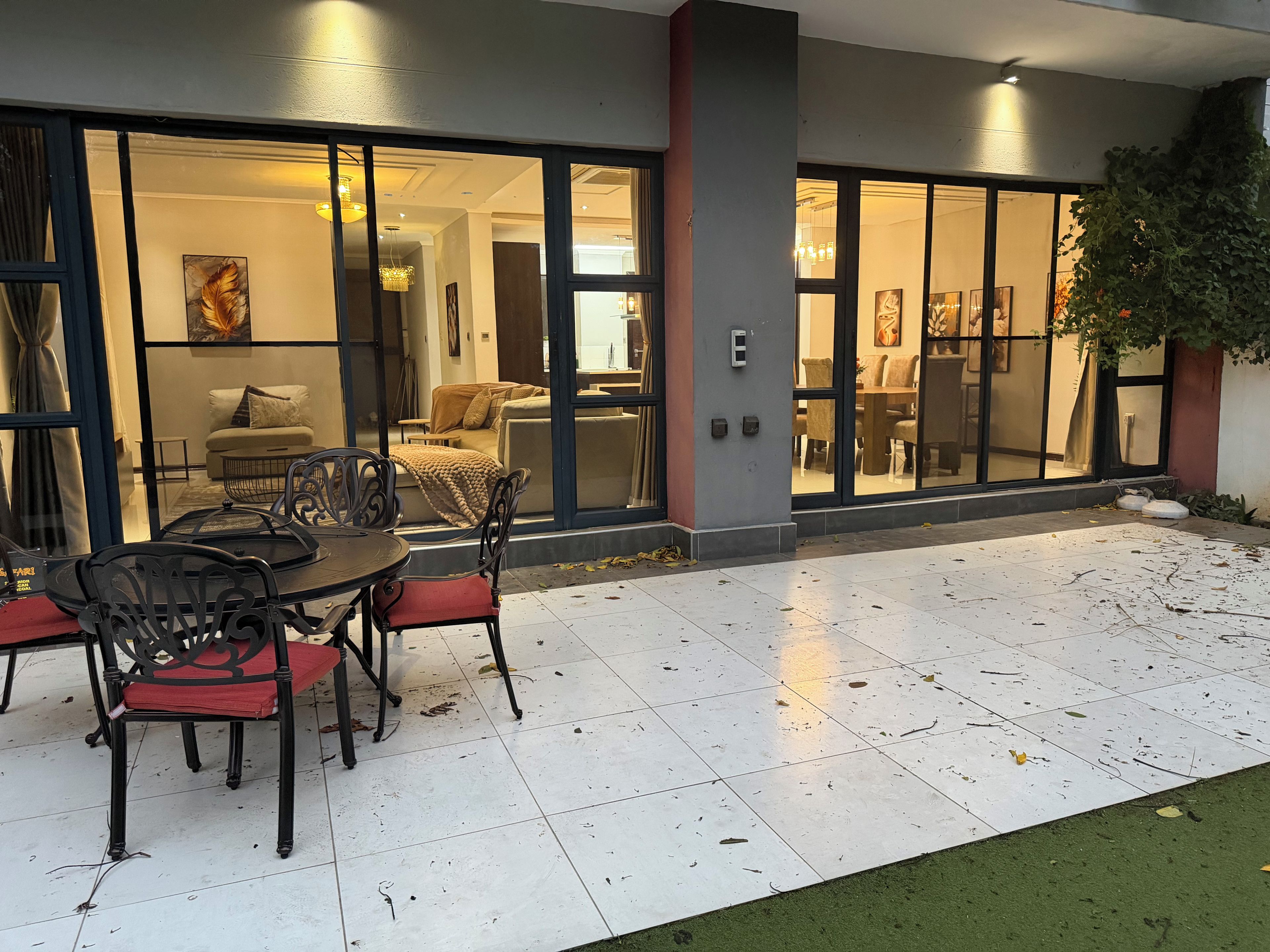 An outdoor patio area with a dining table and chairs, a glass door entrance to a living space, and a small garden area.