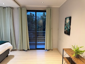 Una camera da letto moderna con un letto, una porta scorrevole in vetro verso un balcone, un quadro al muro e un tavolino con una pianta.
