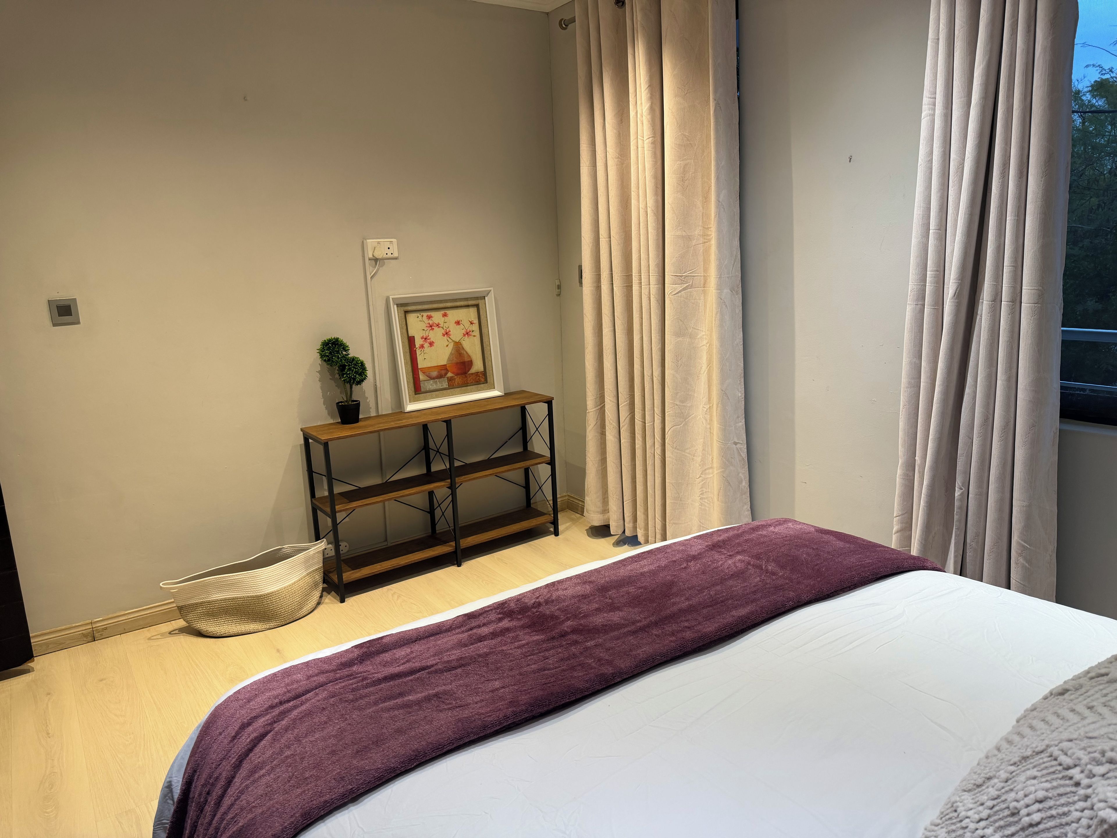 Une chambre à coucher avec un lit, une table de chevet sur laquelle est posée une photo encadrée, une plante en pot et une fenêtre avec des rideaux.