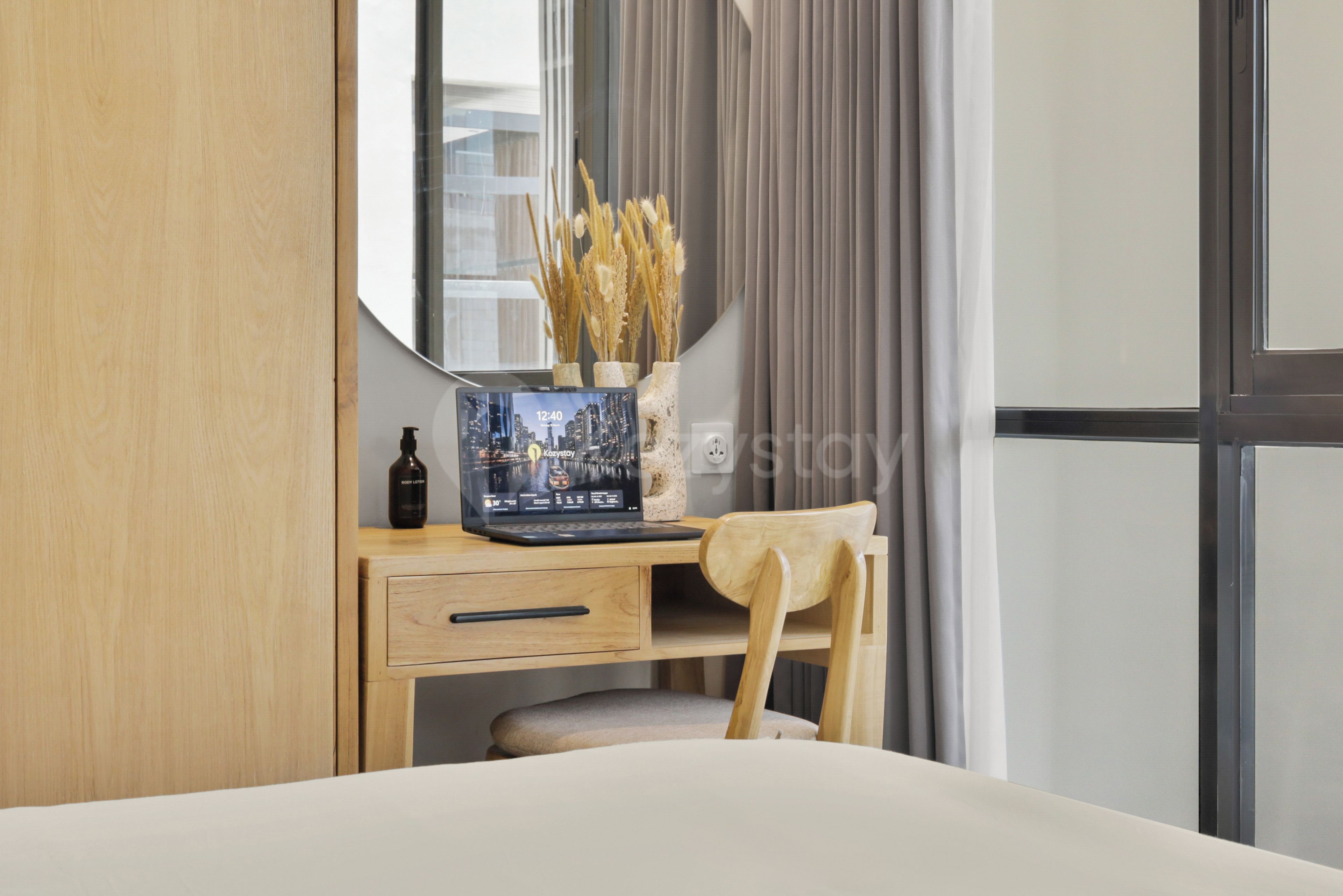 A hotel room with a wooden desk, a laptop, a bottle of body lotion, a vase with dried plants, and a bed with a white comforter.