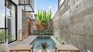 Área de piscina moderna ao ar livre com duas cadeiras de vime e uma mesa pequena, cercada por plantas e um muro de concreto.