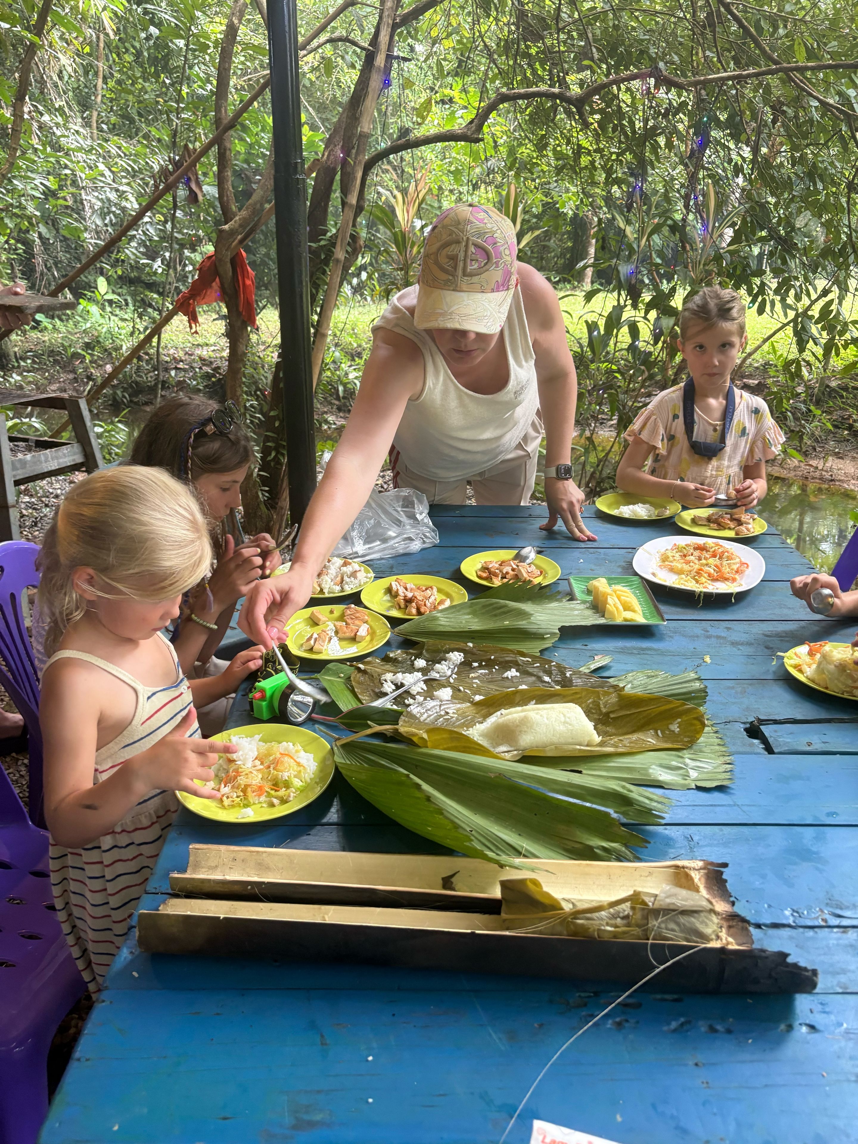 Un groupe de personnes est assis à une table à l’extérieur, avec de la nourriture et des couverts devant elles.