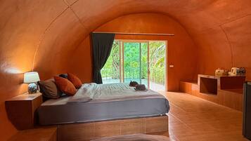 Un cuarto acogedor y curvo con una cama, una mesita de noche, una lámpara y vistas al verdor exterior.