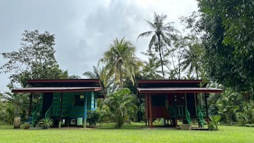 Two small, single-story buildings with green staircases, surrounded by lush greenery and palm trees.