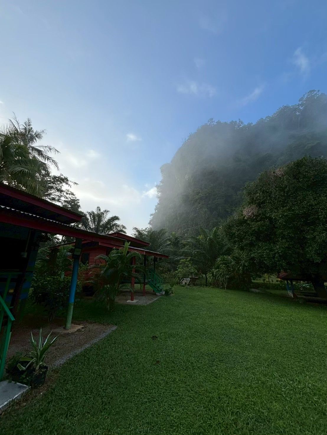 Uma área gramada com um coreto e uma construção de telhado vermelho, cercada por árvores e uma colina nevoenta.