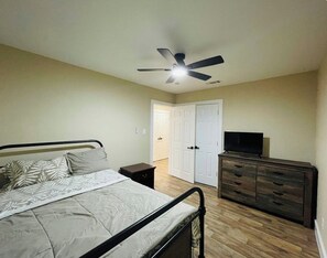 Un dormitorio con cama, cómoda, televisor, ventilador de techo y puerta que da a otra habitación.