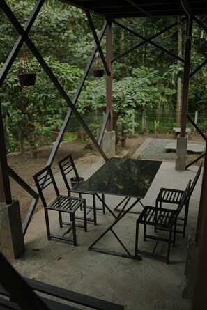 A covered outdoor seating area with a table and chairs, surrounded by greenery.