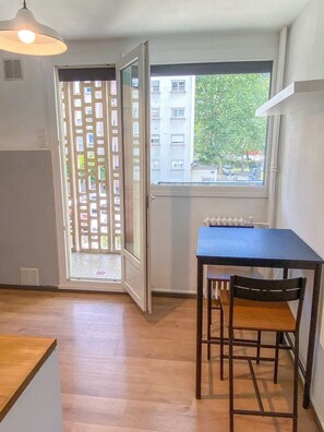 Een kamer met een tafel en stoelen, een radiator, een raam met uitzicht op gebouwen en bomen, en een deur met een lattenhek.