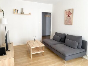 Een moderne woonkamer met een grijze bank, een houten salontafel, een televisie op een houten dressoir, en een boekenkast met boeken en een vaas.