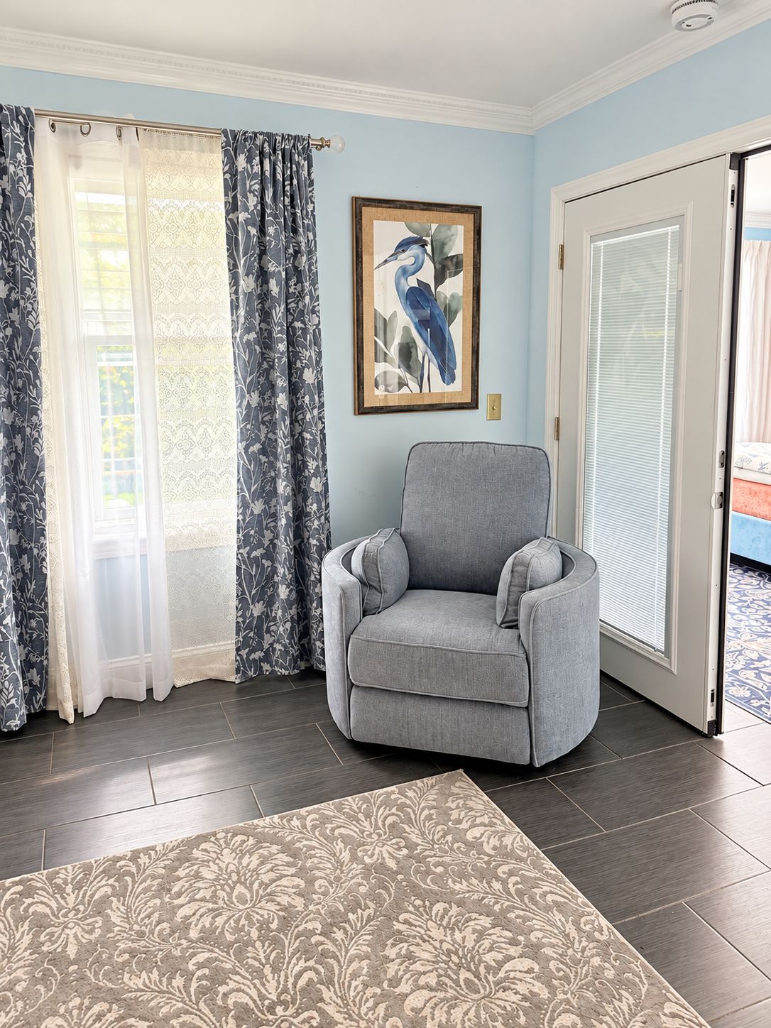 A room with a grey armchair, a patterned rug, a framed bird painting, and a door with blinds.
