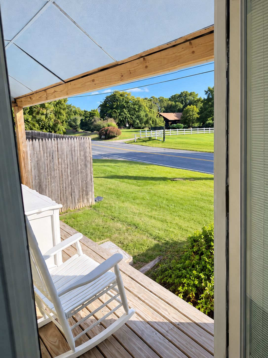 A wooden deck with a white chair, a view of a road, and a grassy area with trees.