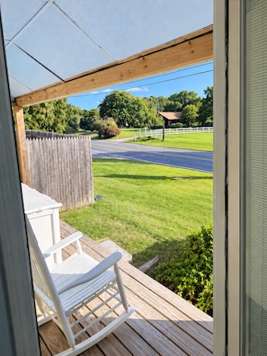 A wooden deck with a white chair, a view of a road, and a grassy area with trees. - Spacious 3-Bedroom Farmhouse in a Charming Equestrian Setting (Tiverton)