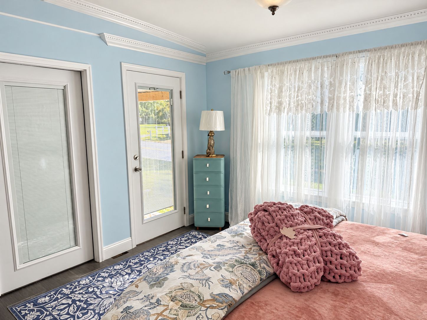 A bedroom with a bed, a dresser, a lamp, and a window with sheer curtains.